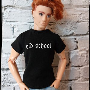 May include: A red-haired doll wearing a black t-shirt with the text "old school" in white. The doll is wearing blue jeans and has a casual, cool look.