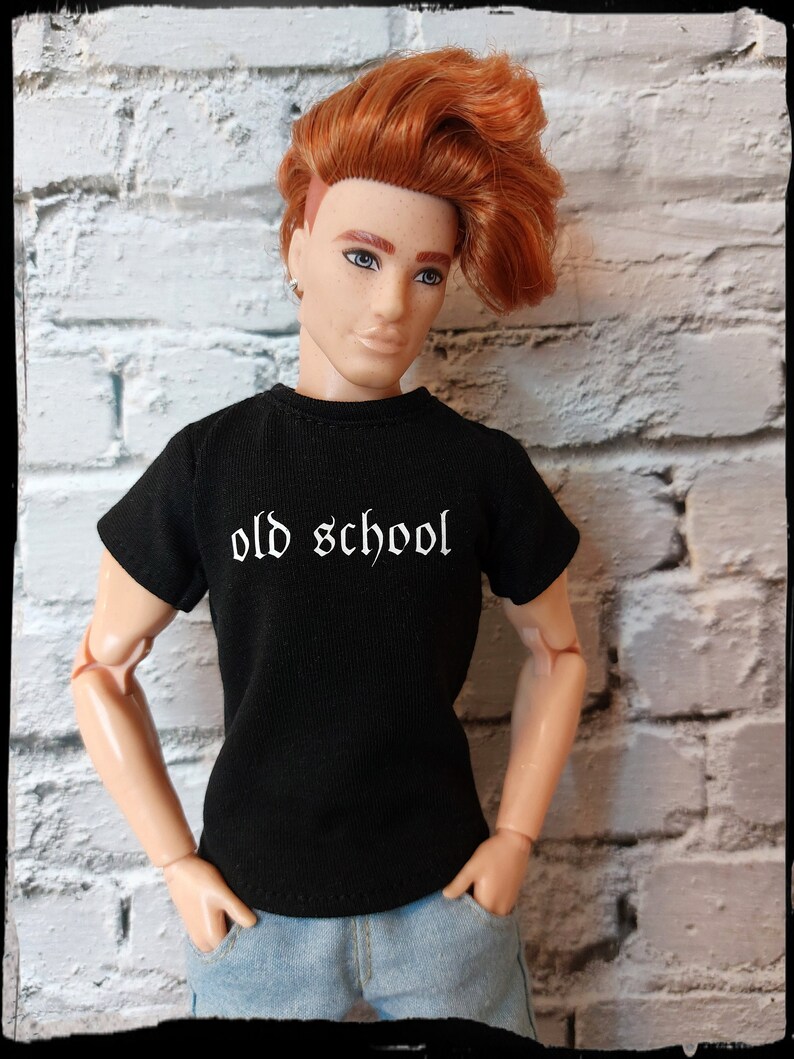 May include: A red-haired doll wearing a black t-shirt with the text "old school" in white. The doll is wearing blue jeans and has a casual, cool look.