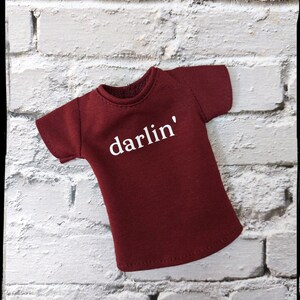 Pu&ograve; includere: Una maglietta bordeaux con la scritta "darlin'" stampata in bianco. La maglietta ha maniche corte e girocollo. La maglietta &egrave; esposta su un muro di mattoni bianchi.