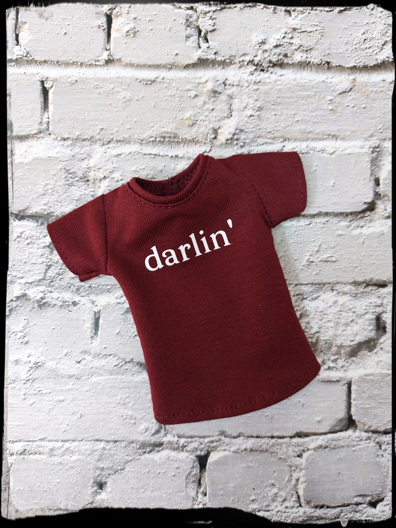 Pu&ograve; includere: Una maglietta bordeaux con la scritta "darlin'" stampata in bianco. La maglietta ha maniche corte e girocollo. La maglietta &egrave; esposta su un muro di mattoni bianchi.