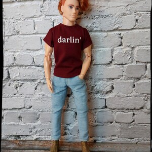 Pu&ograve; includere: Bambola con una maglietta bordeaux con la scritta bianca "darlin'", jeans azzurri e stivali dorati. La bambola ha i capelli rossi ed &egrave; in piedi davanti a un muro di mattoni bianchi.