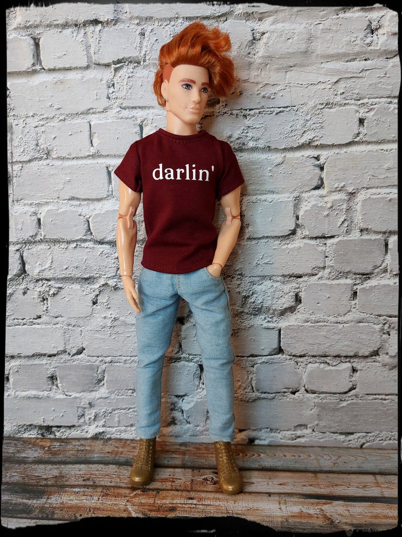 Pu&ograve; includere: Bambola con una maglietta bordeaux con la scritta bianca "darlin'", jeans azzurri e stivali dorati. La bambola ha i capelli rossi ed &egrave; in piedi davanti a un muro di mattoni bianchi.