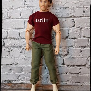 Pu&ograve; includere: Bambola che indossa una maglietta bordeaux con la scritta "darlin'" in bianco, pantaloni verde oliva e stivali color crema. La bambola ha i capelli castani ed &egrave; in posa davanti a un muro di mattoni bianchi.