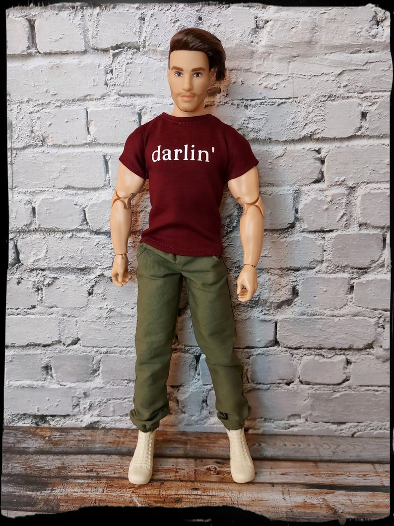 Pu&ograve; includere: Bambola che indossa una maglietta bordeaux con la scritta "darlin'" in bianco, pantaloni verde oliva e stivali color crema. La bambola ha i capelli castani ed &egrave; in posa davanti a un muro di mattoni bianchi.