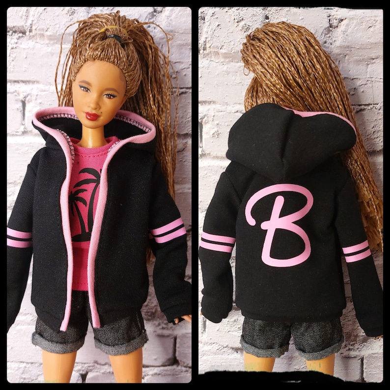 Pode incluir: Camisola de moletom com capuz preta com detalhes em rosa e uma letra "B" rosa nas costas. A camisola tem fecho de correr e capuz com cord&atilde;o.