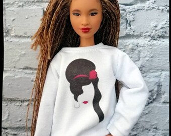 May include: A doll with long brown braids styled in a bun, wearing a white sweatshirt with a black and white graphic of a woman with a red flower in her hair, and blue jeans.