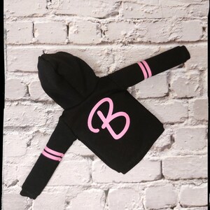 Pode incluir: Camisola de moletom com capuz preta com listras rosa nas mangas e uma grande letra "B" rosa na frente.