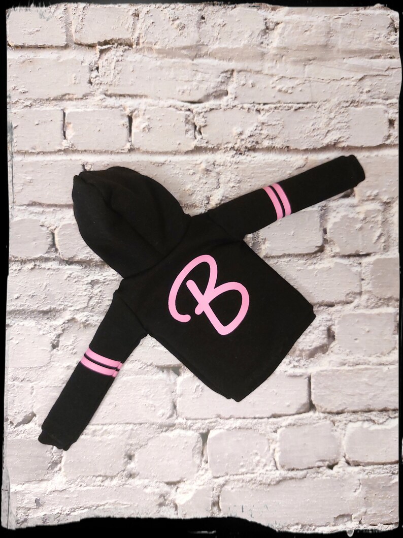 Pode incluir: Camisola de moletom com capuz preta com listras rosa nas mangas e uma grande letra "B" rosa na frente.