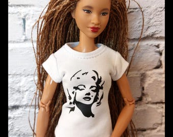 May include: A doll with long brown braids wearing a white t-shirt with a black graphic of Marilyn Monroe and blue jeans. The doll has red lipstick and is posed in front of a white brick wall.