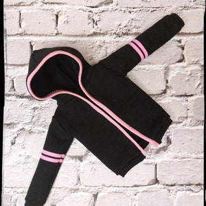 Pode incluir: Camisola de moletom com capuz preta com listras rosa nas mangas e um z&iacute;per rosa. A camisola est&aacute; colocada sobre um fundo de tijolo branco.