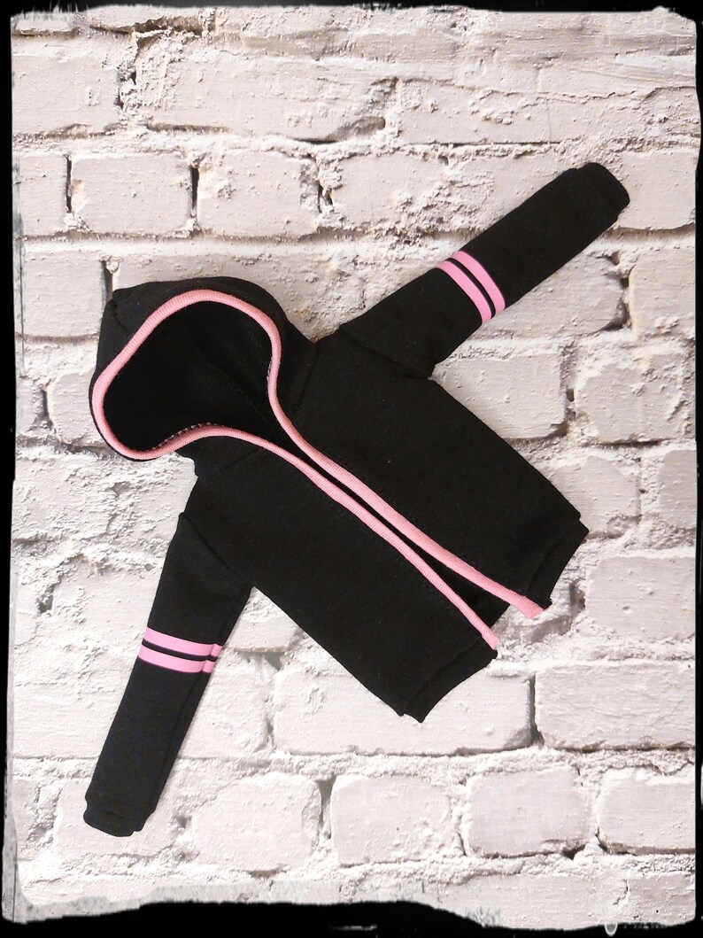 Pode incluir: Camisola de moletom com capuz preta com listras rosa nas mangas e um z&iacute;per rosa. A camisola est&aacute; colocada sobre um fundo de tijolo branco.