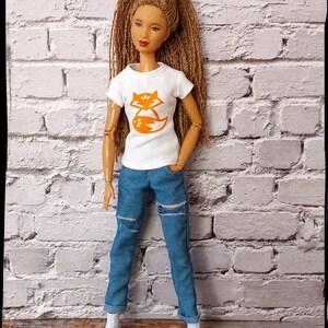 May include: A doll with long brown hair wearing a white t-shirt with an orange fox graphic and blue jeans with distressed details.