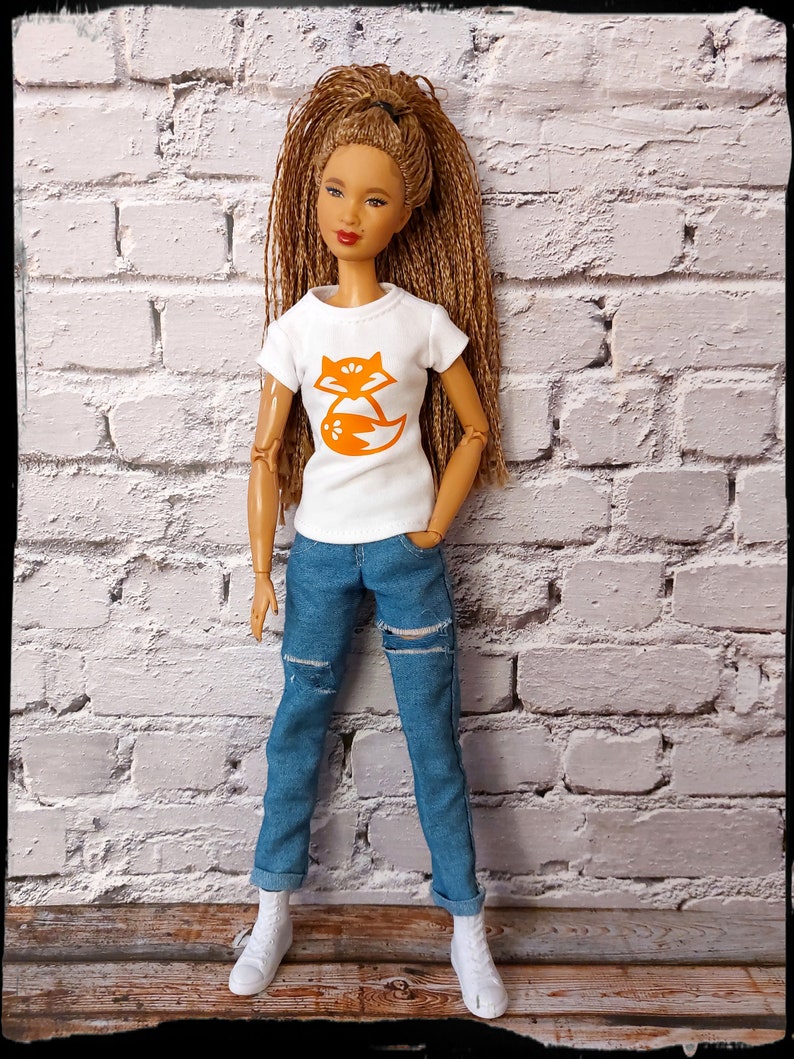 May include: A doll with long brown hair wearing a white t-shirt with an orange fox graphic and blue jeans with distressed details.