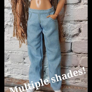 May include: Light blue denim jeans with pockets and a white crop top. The text "Multiple shades!" is displayed in white. The doll is wearing white shoes. The background is a white brick wall.