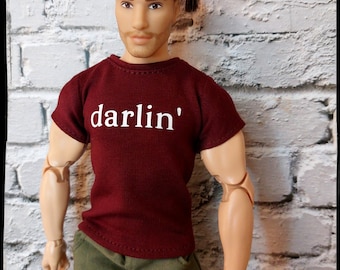 May include: A doll wearing a burgundy t-shirt with the word "darlin'" in white lettering. The doll is also wearing olive green shorts. The doll has brown hair and is posed in front of a white brick wall.