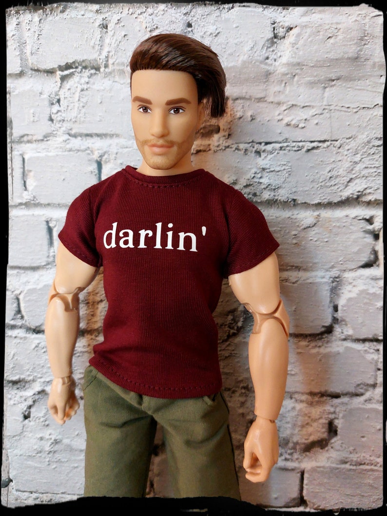 Pu&ograve; includere: Una bambola indossa una maglietta bordeaux con la scritta "darlin'" in lettere bianche. La bambola indossa anche pantaloncini verde oliva. La bambola ha i capelli castani e posa davanti a un muro di mattoni bianchi.