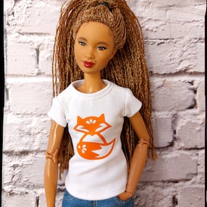May include: A doll with long brown braids wearing a white t-shirt with an orange fox graphic and blue jeans.