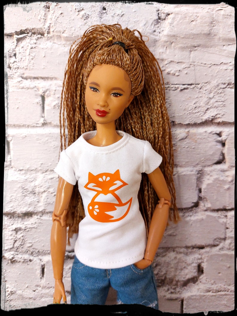 May include: A doll with long brown braids wearing a white t-shirt with an orange fox graphic and blue jeans.