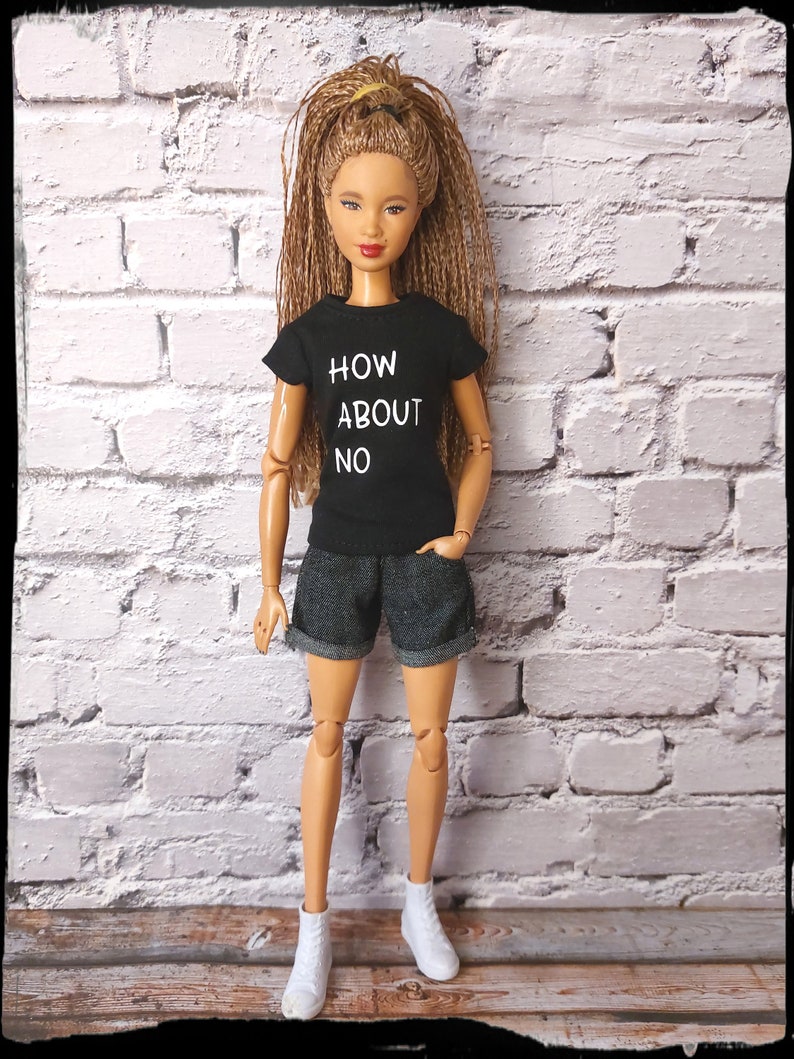 May include: A doll wearing a black t-shirt with the text "HOW ABOUT NO" in white, denim shorts, and white sneakers. The doll has long brown hair and is standing in front of a white brick wall.