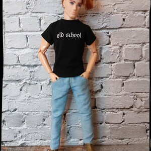 May include: A doll wearing a black t-shirt with the text "old school" in white lettering, blue jeans, and brown shoes. The doll has red hair and is standing in front of a white brick wall.