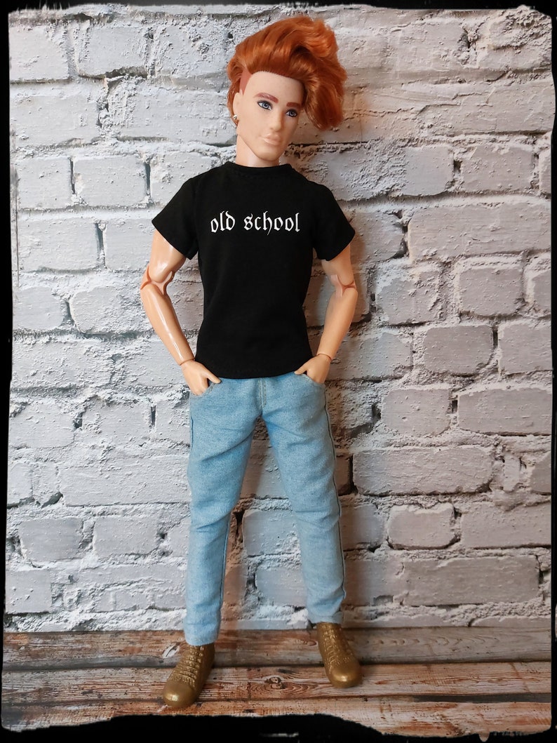 May include: A doll wearing a black t-shirt with the text "old school" in white lettering, blue jeans, and brown shoes. The doll has red hair and is standing in front of a white brick wall.