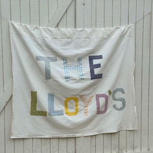 May include: A white fabric banner with the text "THE LLOYD'S" in colorful, patchwork-style letters. The letters are various patterns and colors, including blue, green, yellow, and purple. The banner is hanging against a white wooden background.