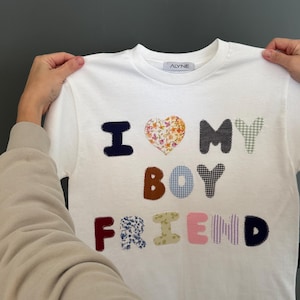 May include: White t-shirt with the words "I love my boyfriend" in colorful patchwork letters. The letters are made of different fabrics with floral, plaid, and striped patterns.