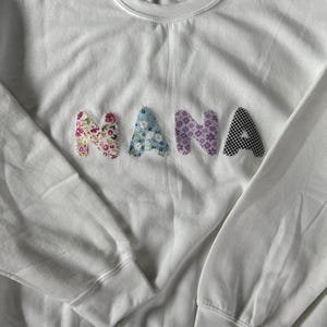 May include: White sweatshirt with the word "MANA" spelled out in colorful fabric patches. Each letter is made of a different patterned fabric.