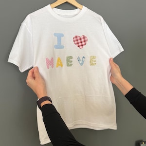 May include: White t-shirt with the phrase "I ❤️ Maeve" in colorful, patchwork-style letters. The heart is red and white checkered, and the name "Maeve" is in pastel colors. The shirt is hanging on a wooden hanger.