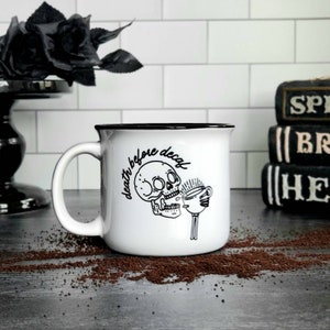 Könnte beinhalten: Weiße Keramik-Tasse mit schwarzem Rand und Henkel, mit einer Totenkopf-Grafik und dem Text "death before decaf". Die Tasse steht auf einer dunklen Oberfläche mit verstreutem Kaffeesatz, mit einer schwarzen Rose und Büchern im Hintergrund.