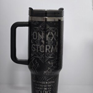May include: Black travel mug with a black handle and lid. The mug features the text "ONYX STORM" and "THERE'S NOWHERE IN EXISTENCE YOU COULD GO THAT I WOULDN'T FIND YOU VIOLENCE ARETIA" in a silver color. The mug has a decorative design.