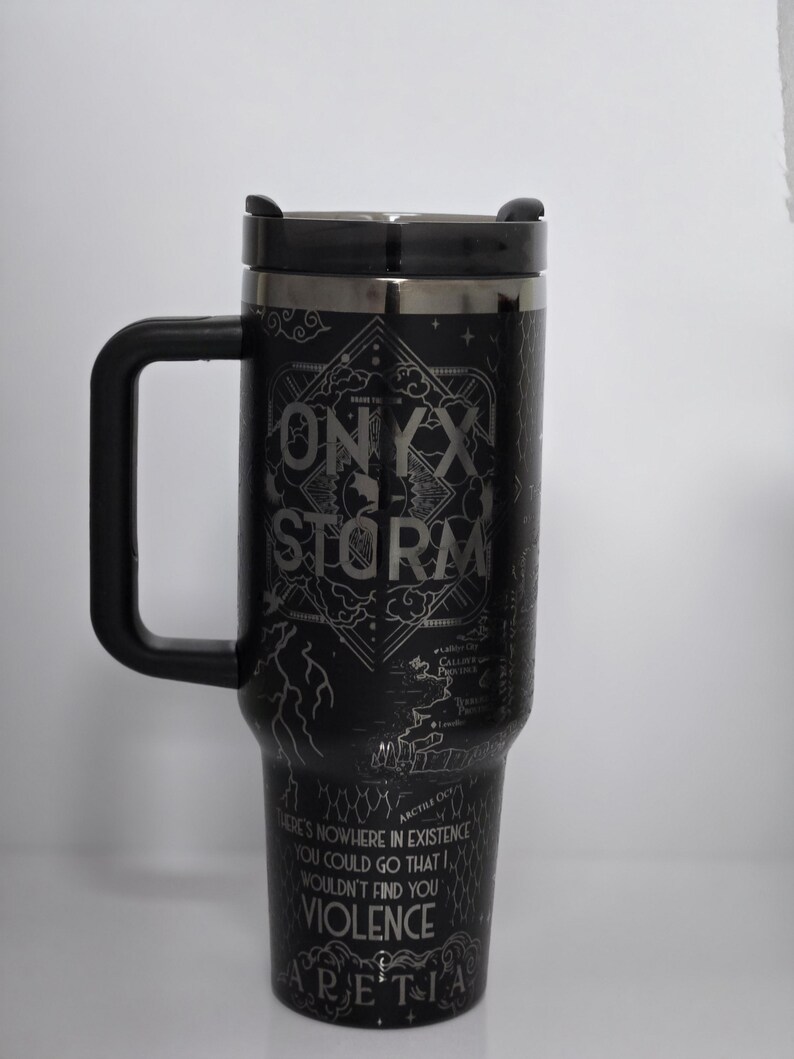 May include: Black travel mug with a black handle and lid. The mug features the text "ONYX STORM" and "THERE'S NOWHERE IN EXISTENCE YOU COULD GO THAT I WOULDN'T FIND YOU VIOLENCE ARETIA" in a silver color. The mug has a decorative design.