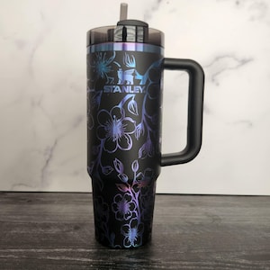 Könnte beinhalten: Schwarze Reise-Thermoskanne aus Edelstahl mit Blumenmuster und schwarzem Griff. Die Kanne hat einen schwarzen Deckel mit einem Strohhalmloch und das Stanley-Logo ist auf der Seite aufgedruckt.