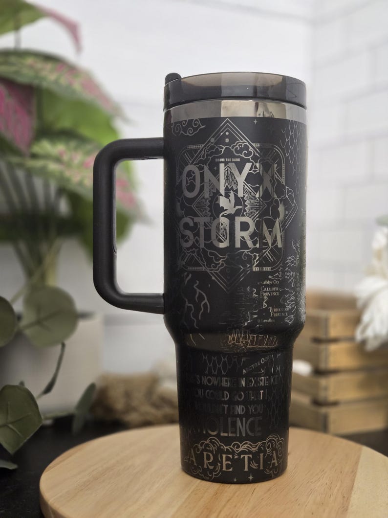 May include: Black travel mug with a handle, featuring the text "ONYX STORM" and other script. The mug has a black lid and a silver rim. The mug is on a wooden surface, with plants in the background.
