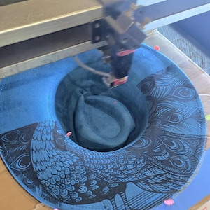 May include: A blue fedora hat featuring a peacock design, in the process of being laser cut. The hat has a dark blue brim and a lighter blue crown. The peacock design is black.