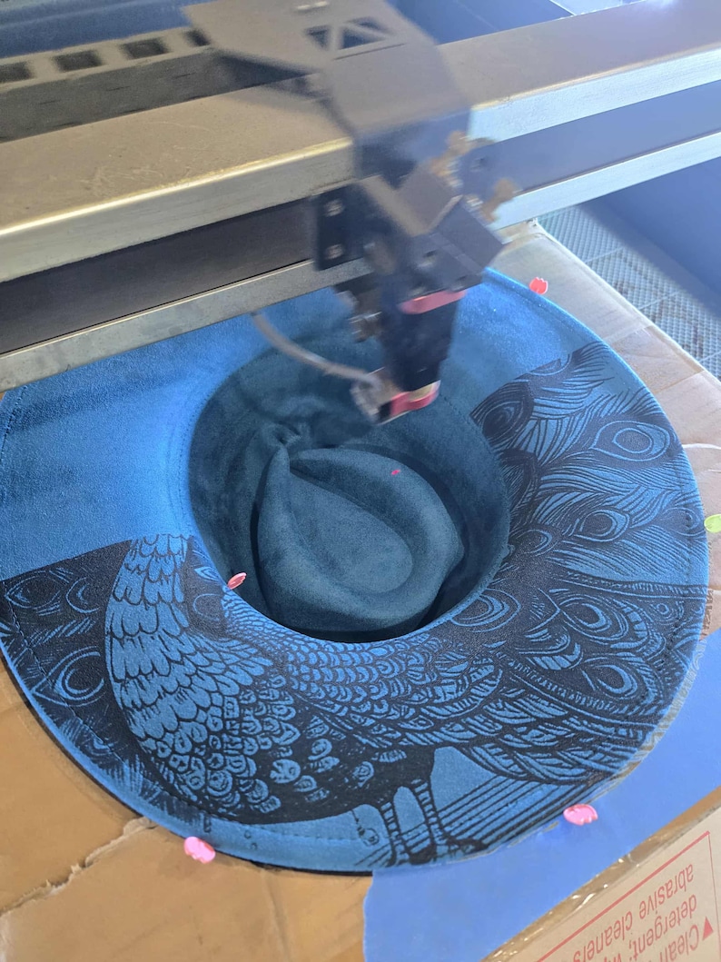 May include: A blue fedora hat featuring a peacock design, in the process of being laser cut. The hat has a dark blue brim and a lighter blue crown. The peacock design is black.