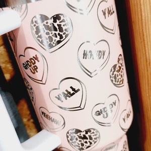 May include: Pink stainless steel tumbler with a clear lid and a straw. The tumbler has a pattern of silver hearts with various sayings, including "Giddy Up", "Y'all", and "Howdy".