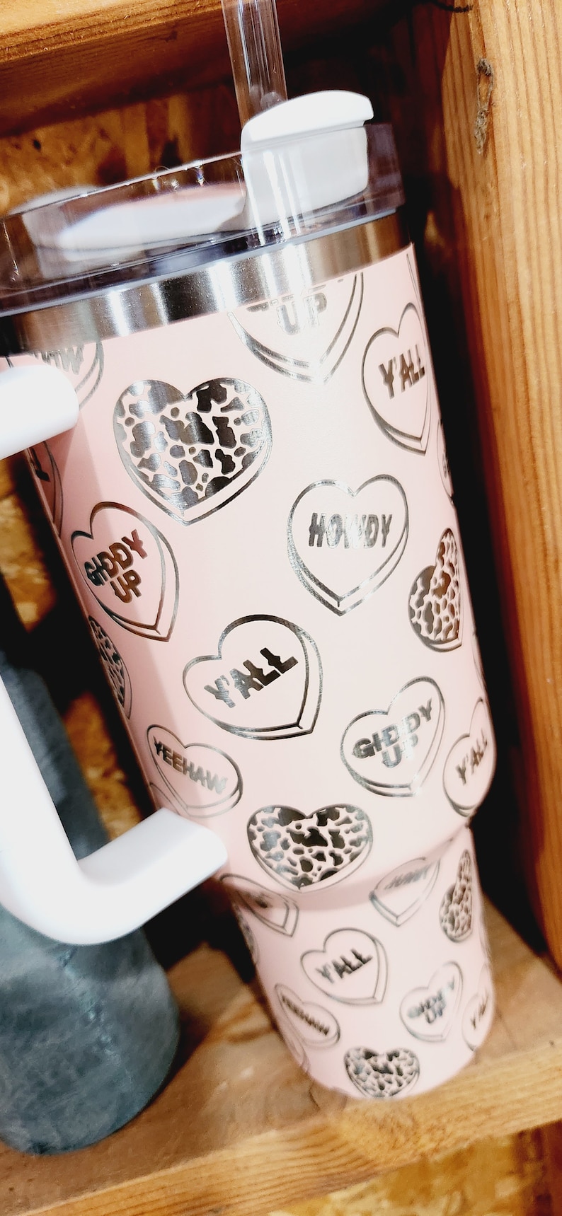 May include: Pink stainless steel tumbler with a clear lid and a straw. The tumbler has a pattern of silver hearts with various sayings, including "Giddy Up", "Y'all", and "Howdy".