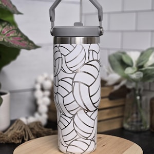 Puede incluir: Un vaso térmico blanco con asa y tapa grises. El vaso está cubierto con un estampado de balones de voleibol en blanco y negro. Está colocado sobre una tabla de cortar de madera.