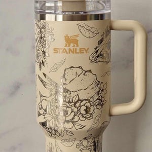 Puede incluir: Una taza de viaje de acero inoxidable de color crema con un dise&ntilde;o floral negro y dorado. La taza tiene un asa y una tapa. La taza tiene la palabra "STANLEY" impresa en el lateral.