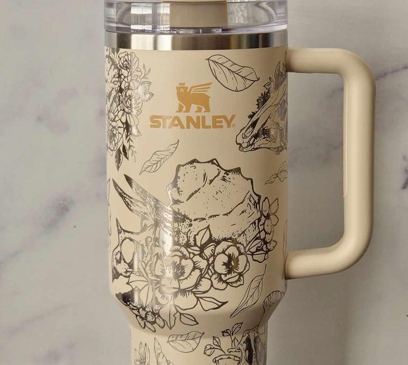 Puede incluir: Una taza de viaje de acero inoxidable de color crema con un dise&ntilde;o floral negro y dorado. La taza tiene un asa y una tapa. La taza tiene la palabra "STANLEY" impresa en el lateral.