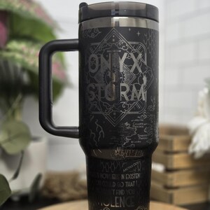 May include: Black travel mug with a handle and a black lid. The mug features the words "ONYX STORM" in a decorative font, along with other text and a map-like design. The bottom of the mug has the word "ARETIA".