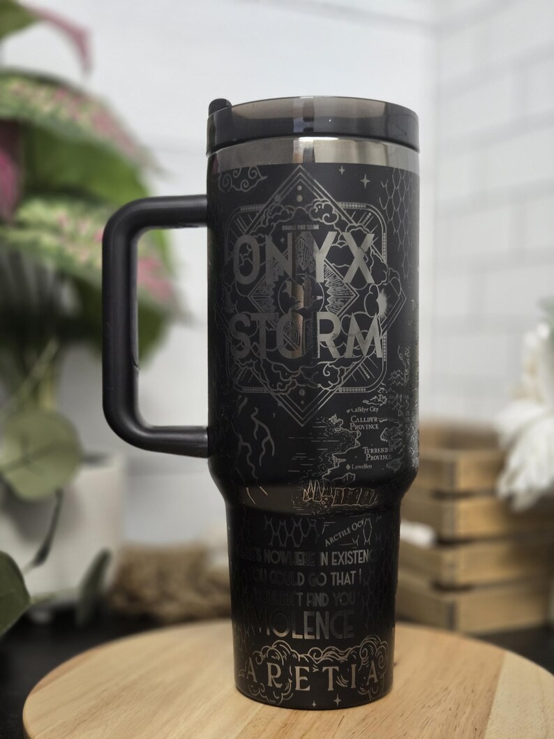 May include: Black travel mug with a handle and a black lid. The mug features the words "ONYX STORM" in a decorative font, along with other text and a map-like design. The bottom of the mug has the word "ARETIA".