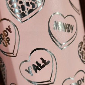 May include: Pink stainless steel tumbler with a silver heart pattern. The hearts have different sayings on them, including "Howdy", "Giddy Up", "Y'all", and "Yeehaw".