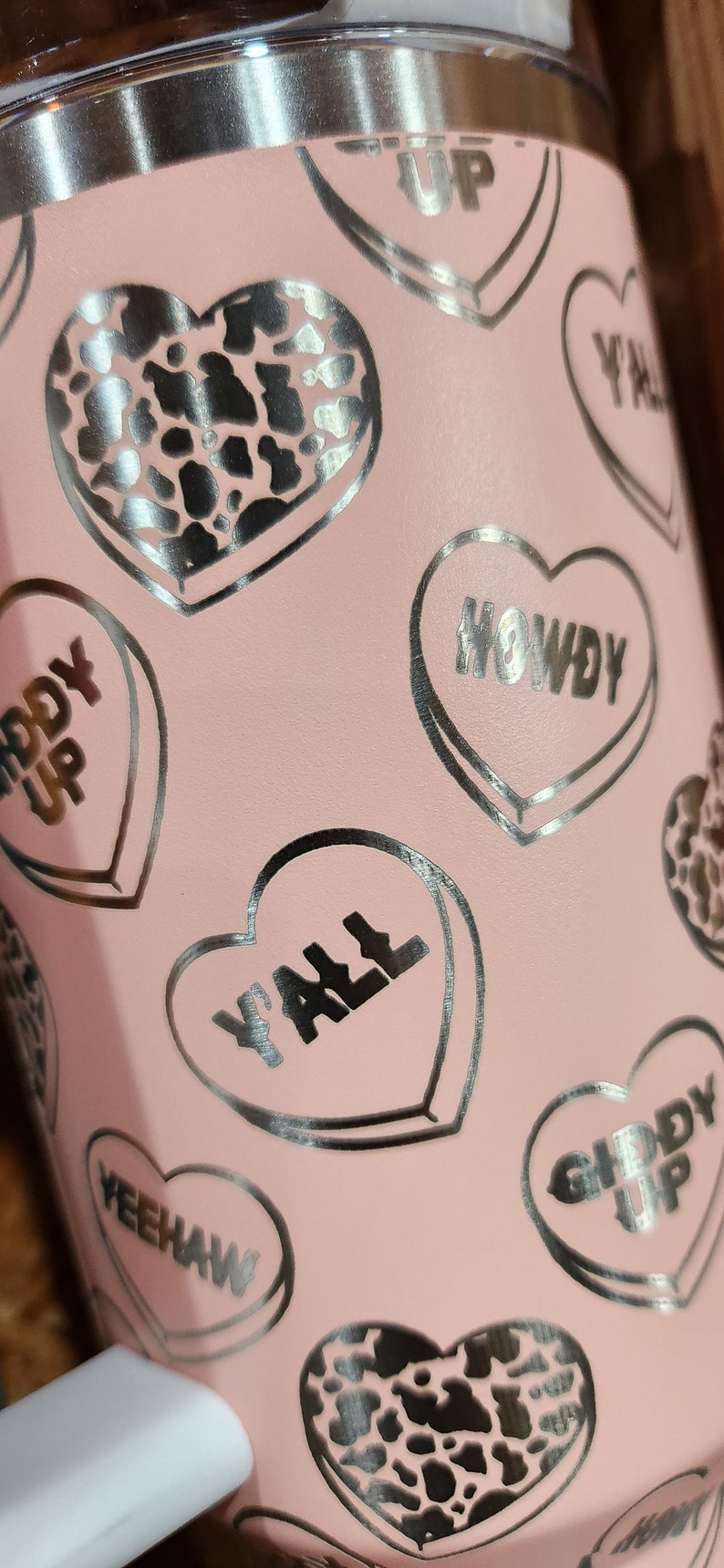 May include: Pink stainless steel tumbler with a silver heart pattern. The hearts have different sayings on them, including "Howdy", "Giddy Up", "Y'all", and "Yeehaw".