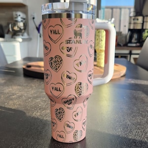 Peut inclure: Un gobelet isotherme en acier inoxydable rose avec un couvercle et une paille dorés. Le gobelet est orné d'un motif de cœurs dorés avec différentes inscriptions, notamment "Y'all", "Giddy Up" et "Yeehaw". Le gobelet porte également le logo Stanley et les mots "Get Up".