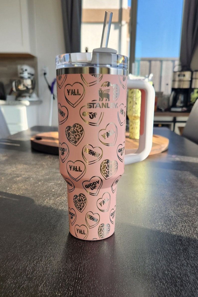 May include: A pink stainless steel tumbler with a gold lid and straw. The tumbler has a pattern of gold hearts with various sayings, including "Y'all", "Giddy Up", and "Yeehaw". The tumbler also has the Stanley logo and the words "Get Up".