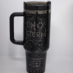 May include: Black travel mug with a black handle and lid. The mug has the words "ONYX STORM" in a silver font, along with other text and a map design. The mug is cylindrical and tapers slightly.