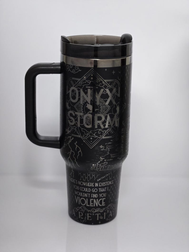 May include: Black travel mug with a black handle and lid. The mug has the words "ONYX STORM" in a silver font, along with other text and a map design. The mug is cylindrical and tapers slightly.