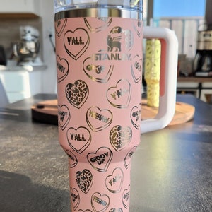 May include: Pink stainless steel tumbler with a white lid and a straw. The tumbler has a gold print of hearts with various sayings, including "Y'all", "Giddy Up", and "Yeehaw". The tumbler also has the Stanley logo printed on it.